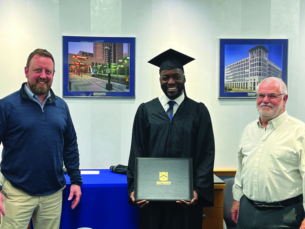 Graduate proudly holds diploma flanked by two men in an office.