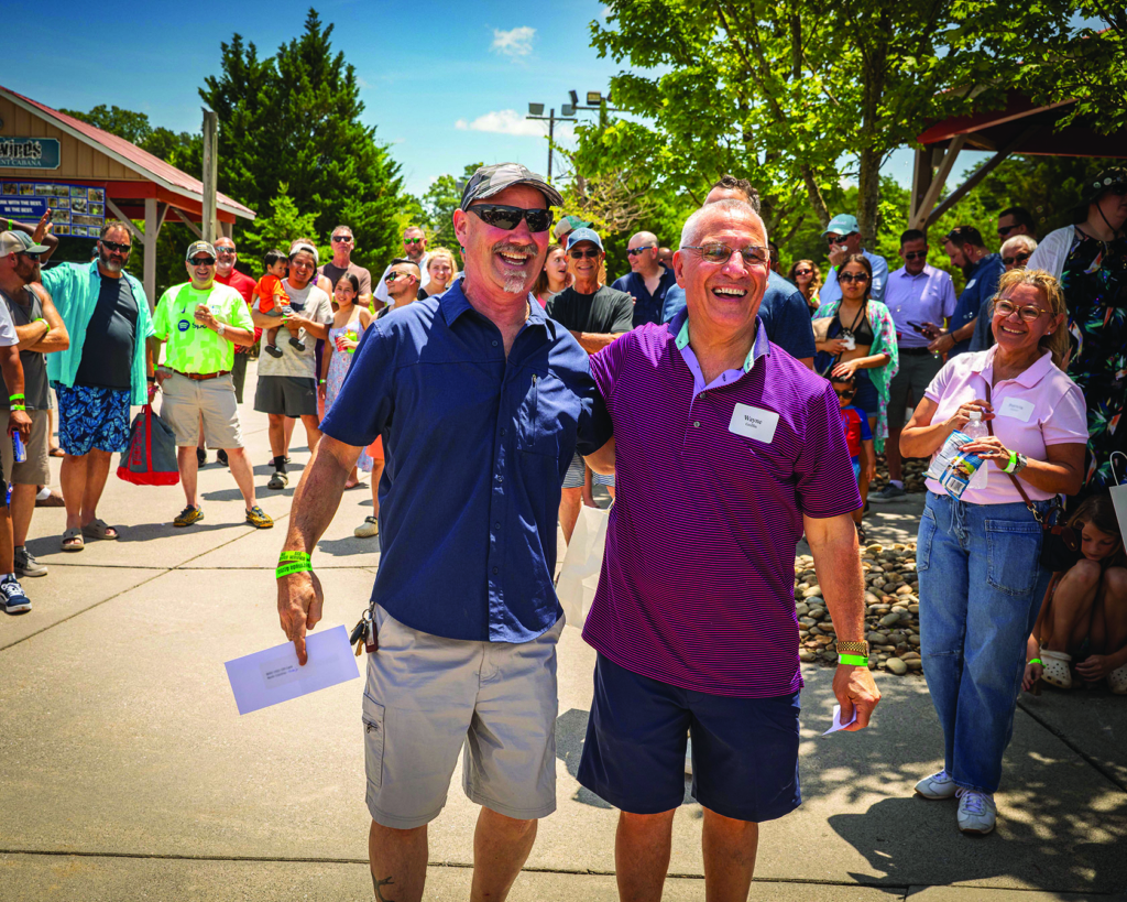 Two men smiling and walking together at an outdoor event on a sunny day.