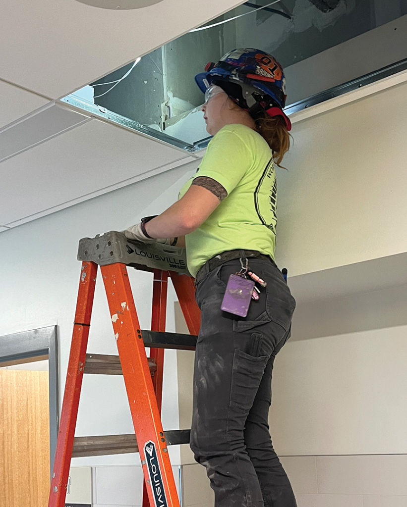 Worker on ladder wearing safety gear and gloves indoors.